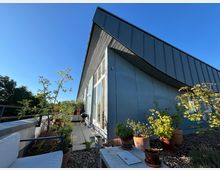 Metallverkleidetes Gebäude mit geneigtem Dach, angrenzend an eine Terrasse mit Pflanzkübeln, Sitzgelegenheiten und einem Grill, umgeben von Bäumen und klarem Himmel im Hintergrund.