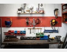Werkstatt mit einer roten Lochwand, an der Werkzeuge wie Schraubenschlüssel, Schutzbrillen und Gehörschutz hängen. Darunter befinden sich eine Werkbank mit Schraubstock, Ablagekästen für Schrauben und Muttern sowie ein Akkuschrauber.