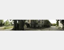 Ein Innenhof mit Kopfsteinpflaster, umgeben von historischen Gebäuden, darunter ein grünes Haus mit Satteldach und ein graues Gebäude mit Außentreppe. Im Hintergrund sind kleine Bäume, ein Zaun und Teile weiterer Bauwerke zu sehen.