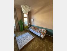 Ein schlicht eingerichtetes Zimmer mit einer Liege, die eine bunte Blumendecke hat, einer Matte auf dem Teppichboden, einem grünen Kaktus-Dekor, einem gelben Stuhl und einer wolkenförmigen Deckenleuchte. Zwei große Fenster mit beigefarbenen Vorhängen lassen Tageslicht herein.