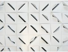 Nahaufnahme einer modernen, weißen Wand- oder Deckenverkleidung aus quadratischen Paneelen mit gefalteten, dreidimensionalen Dreiecksformen. Zwischen den gefalteten Flächen verlaufen diagonal angeordnete dunkle Schlitze, die ein geometrisches Muster bilden.
