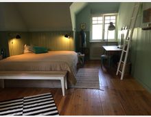 Schlafzimmer mit Holzboden und grün gestrichenen Holzwänden. Ein Doppelbett mit beiger Tagesdecke, eine Leiter an der Wand, ein kleiner Tisch mit Stuhl und eine große Fensterfront prägen den Raum.