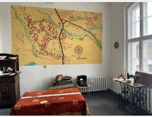 Ein musealer Raum mit einer historischen Karte von Vierraden an der Wand. Im Vordergrund stehen ein Tisch mit roter Decke, antike Möbel, eine Schreibmaschine und eine alte Nähmaschine, während rechts ein großes Fenster natürliches Licht einlässt.