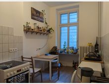 Kleine Küche mit weißen Wänden, Fliesen an der Wand nahe dem Herd und einem Holzfußboden. Ein Esstisch mit zwei Stühlen steht in der Mitte, dahinter ein Fenster mit Fensterbank, auf der Küchenutensilien und Obst stehen. Ein Regal mit Gewürzen und Pflanzen hängt an der Wand, während rechts Spüle, Haushaltsgeräte und ein Heizkörper zu sehen sind.