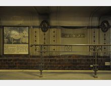 Untergrundbahnhof-Station „Breitenbachplatz“ mit Jugendstil-Elementen, bestehend aus braunen Wandfliesen, einem dekorativen Geländer und Wandverzierungen. Auf der linken Seite ein historisches Foto mit Texttafel, rechts das Stationsschild.