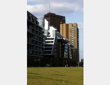 Moderne Stadtansicht mit mehreren Gebäuden, darunter Glas- und Metallfassaden sowie ein brauner Hochhausturm im Hintergrund. Im Vordergrund erstreckt sich eine große, offene Grünfläche.