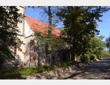 Ansicht einer Kirche mit roten Ziegeldächern und verputzten sowie teils aus Backstein bestehenden Wänden, umgeben von Bäumen. Rechts verläuft eine gepflasterte Straße mit einer Steinmauer am Rand.