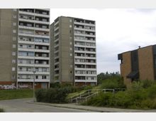 Zwei mehrstöckige Wohnblöcke in Plattenbauweise und ein modernes Gebäude aus Ziegelstein mit einem Kreuz auf dem Dach. Im Vordergrund befinden sich Grünflächen, Büsche und ein Fußweg mit Geländer.