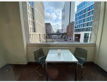 Kleiner Balkon mit weißem Tisch, zwei grauen Stühlen und einer Lichterkette an der Balkonbrüstung. Im Hintergrund ist eine Stadtansicht mit modernen Bürogebäuden und einer Straße zu sehen.