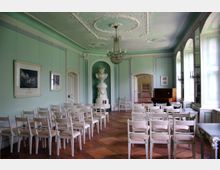 Innenraum eines historischen Saals mit mintgrünen Wänden und weißem Stuck an der Decke. Der Raum ist mit weißen Stühlen in Reihen, einem Klavier, großen Fenstern mit viel Tageslicht und einem großen Kachelofen dekoriert.