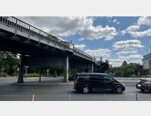 Straßenszene mit einer erhöhten Stahlbrücke, die diagonal über die Straße verläuft. Darunter befinden sich Ampeln, Straßenverkehr und Gehwege. Im Hintergrund sind Bäume sowie die Spitze eines roten Gebäudeturms zu sehen, und am Himmel befinden sich einige Wolken.