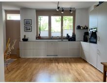 Helle, moderne Küche mit weißen Einbauschränken, Holzfußboden und einem großen Fenster mit Blick ins Grüne. Links eine Eingangstür, darüber kleine Wandbilder, rechts ein eingebauter Backofen und Küchenutensilien auf der Arbeitsfläche.