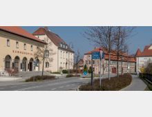 Straßenszene in einer kleinen Stadt mit mehreren historischen Gebäuden, darunter die „Kammerspiele“ mit Rundbogenarkaden. Links ein sandfarbenes Wohnhaus mit roten Dachziegeln, rechts eine Fachwerkfassade und Gehwege mit jungen, unbelaubten Bäumen.