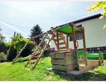 Ein Spielplatz mit einem hölzernen Klettergerüst, einer Rutsche und einem angebauten Sandkasten, umgeben von Rasen und Bäumen; im Hintergrund ein cremefarbenes Gebäude mit kleinen Fenstern.