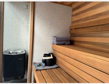 Innenraum einer modernen Sauna mit Holzbänken und holzverkleideten Wänden. Auf der linken Seite befindet sich ein Saunaofen, auf der Bank liegen gerollte Handtücher sowie ein Eimer mit Wasser und Duftölen.