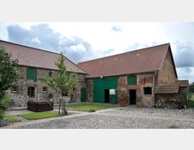 Ein Bauernhof mit traditionellen Backsteingebäuden, roten Ziegeldächern und grünen Holztüren. Der Hof hat einen gepflasterten Innenhof mit kleinen Grünflächen, Bäumen und Holzstapeln an der Seite.