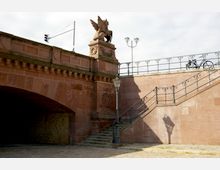 Sandsteinbrücke mit dekorativen Elementen, darunter ein geflügeltes Fabelwesen als Skulptur auf einem Sockel. Eine Treppe mit schmiedeeisernem Geländer führt zur Oberseite der Brücke, daneben eine Straßenlaterne und ein Fahrrad auf dem Gehweg.