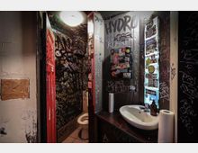 Öffentliche Toilettenräume mit stark bemalten und beschrifteten Wänden, teils mit Graffiti und Aufklebern bedeckt. Zu sehen sind eine Toilette, ein Waschbecken mit Seifenspender und Papierrollen sowie eine rote Tür.