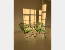 Zwei grüne Stühle mit drahtähnlichen Sitzflächen stehen in einem minimalistischen Raum mit beigem Boden. Im Hintergrund fällt warmes, goldenes Licht durch ein Fenster und erzeugt rechteckige Schattenmuster an der Wand.
