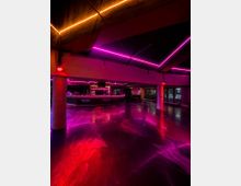 Innenraum eines modernen Nachtclubs mit geschwungener Bar und spiegelglänzendem Boden, der pinke und rote Neonlichter reflektiert. Sichtbare Deckenbalken, LED-Lichtstreifen und Stützsäulen rahmen den offenen Raum mit großen Fensterfronten auf der rechten Seite.