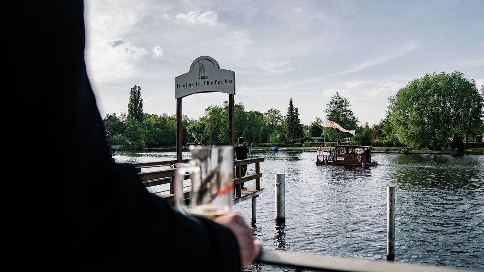 Blick auf eine Holzplattform am Wasser mit dem Schild „Freiheit Fünfzehn“. Im Hintergrund ein kleines Boot auf einem Fluss, umgeben von Bäumen unter einem klaren Himmel.