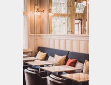 Innenraum eines Cafés mit gepolsterten Sitzbänken entlang einer Wandvertäfelung in hellem Holz. Davor stehen marmorne Tische, schwarze Ledersessel und mehrere farbige Kissen als Dekoration; an der Wand befindet sich ein großer Spiegel, der Licht reflektiert.