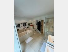 Modernes Badezimmer mit hellen Fliesen, einer Badewanne, einer Dusche mit Glastür, zwei Waschbecken und einem Handtuchheizkörper. An den Wänden hängen gerahmte Bilder, und es sind Pflegeprodukte sowie Handtücher sichtbar.