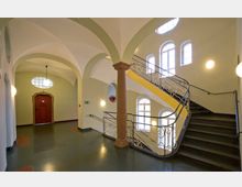 Treppenhaus in einem historischen Gebäude mit gewölbter Decke, einer runden Säule, geschwungenem Metallgeländer und mehreren hohen Fenstern, die den Raum mit natürlichem Licht erhellen. Im Hintergrund befindet sich eine rote Holztür mit einem steinernen Türrahmen.