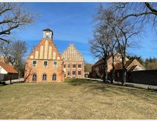 Historisches Backsteingebäude mit Stufengiebeln auf einem offenen, grasbewachsenen Platz, umgeben von weiteren Backsteingebäuden und kahlen Bäumen. Der klare blaue Himmel deutet auf sonniges Wetter hin.
