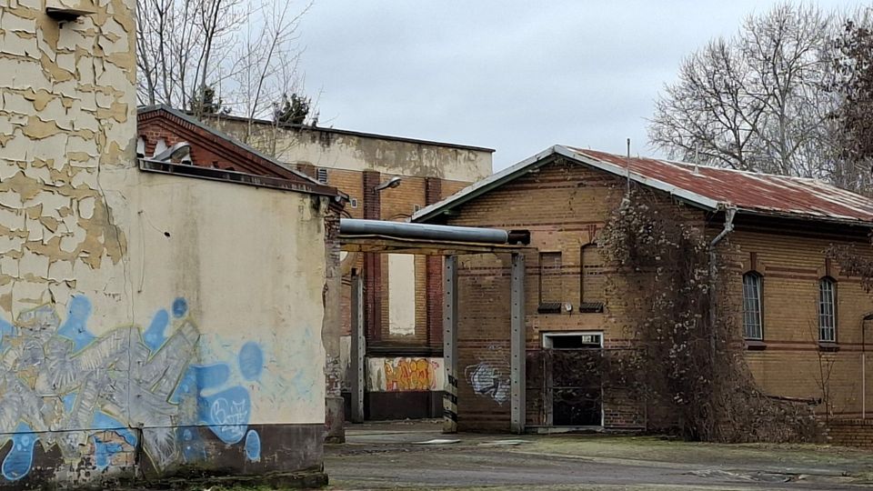 Verlassener Industriehof mit teilweise verputzten Backsteingebäuden, abblätternder Fassade und Graffiti an einer Seitenwand. Im Hintergrund stehen flache Hallen mit rostigem Blechdach, schmalen Fenstern, überwucherten Sträuchern und kahlen Bäumen.