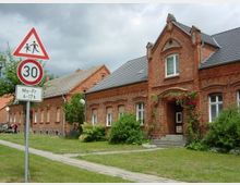 Rote Backsteinhäuser mit Satteldächern stehen entlang einer gepflasterten Gehwegseite, umgeben von Grünflächen und Sträuchern. Im Vordergrund ein Verkehrsschild mit einer Geschwindigkeitsbegrenzung von 30 km/h und einem Hinweis auf Schulzeit (Mo-Fr 6-17 Uhr).