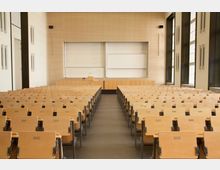 Ein großer, moderner Hörsaal mit mehreren Reihen aus hellen Holzsitzen, einem zentralen Mittelgang und einem Rednerpult vor zwei großen weißen Tafeln an der Vorderwand. Rechts sind hohe Fenster, die Tageslicht in den Raum lassen, und die Wände sind mit Holzpaneelen verkleidet.