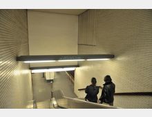 Treppenabgang in einer U-Bahn-Station mit beige gefliesten Wänden. Zwei Personen stehen auf einer Rolltreppe, und darüber ist eine beleuchtete Deckenlampe mit Graffiti zu sehen.