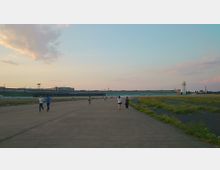 Weitläufiges Gelände eines ehemaligen Flughafens mit einer großen, flachen Landebahn im Vordergrund, gesäumt von Wiesen. Im Hintergrund befindet sich ein langgestrecktes Gebäude mit flachem Dach und ein markanter Turm, während Spaziergänger und Radfahrer die Szene beleben.