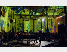 Eine Veranstaltungshalle mit industriellem Charme, beleuchtet durch großflächige, bunte Projektionen in Gelb- und Blautönen. Im Vordergrund steht ein Flügel auf einer Bühne, umgeben von Publikum; zwei Personen verbeugen sich vor den Zuschauenden.