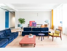 Ein modernes Wohnzimmer mit hellen Wänden und großen Fenstern, die durch weiße und gelbe Vorhänge eingerahmt sind. Im Vordergrund stehen dunkelblaue Sofas, ein roter Couchtisch und ein gelber Sessel, ergänzt durch dekorative Lampen, eine Topfpflanze und minimalistische Kunstwerke im Hintergrund.