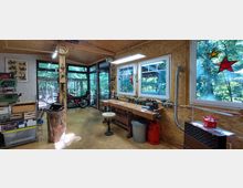 Werkstatt mit Holzwänden und großen Fenstern, die in einen grünen Garten blicken. Ein Arbeitstisch mit Werkzeugen, ein Motorrad, eine Gasflasche und diverse Arbeitsgeräte prägen den Raum.