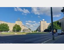 Weitläufiger Platz mit einem zentralen Springbrunnen, umgeben von neoklassizistischen Gebäuden mit heller Fassade. Im Vordergrund eine mehrspurige Straße, im Hintergrund Bäume und blauer Himmel mit vereinzelten Wolken.