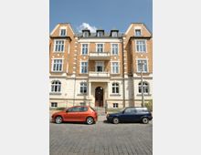 Mehrstöckiges historisches Wohngebäude mit einer Fassade aus rotem Backstein und beigem Putz, großen Fenstern und einem zentralen Eingang mit Säulen. Vor dem Gebäude stehen zwei geparkte Autos, eines orangefarben und eines blau, auf einer gepflasterten Straße.