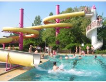 Freibad mit einer großen gelben Wasserrutsche, die von pinken Säulen gestützt wird, und einer weißen Wendeltreppe, auf der Menschen anstehen. Im Vordergrund ein Becken mit mehreren Personen, teils schwimmend, teils spielend, umgeben von grünem Buschwerk und Bäumen.