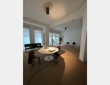 Großer, heller Raum mit weißen Wänden und hohen Fenstern, die mit halbtransparenten Rollos bedeckt sind. Im Vordergrund ein runder Tisch mit Arbeitsutensilien, umgeben von Stühlen, im Hintergrund eine Leiter, ein Sofa und mehrere lose Stühle, auf braunem Teppichboden.