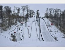 Schnee bedeckte Skisprunganlage mit mehreren Anlaufbahnen unterschiedlicher Größe, umgeben von Bäumen und einem hügeligen, verschneiten Waldgelände. Im Hintergrund ist ein Gebäude am Waldrand sichtbar.
