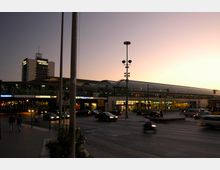 Bahnhofsumgebung bei Dämmerung mit beleuchtetem Glasdach des Spandauer Bahnhofs im Hintergrund, mehreren Laternen und dem Hochhaus der Spandau Arcaden. Im Vordergrund ist eine belebte Kreuzung mit Autos, Rollern und Fußgängern zu sehen.