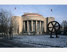 Außenansicht der Volksbühne in Berlin, einem historischen Theatergebäude mit breiter Fassade, Säulen und dem Schriftzug "VOLKSBÜHNE". Vor dem Gebäude stehen kahle Bäume, eine große Metallskulptur in Form eines Rades und ein Reisebus; der Boden ist mit leichtem Schnee bedeckt.