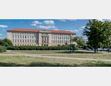 Ein großes, mehrstöckiges Gebäude mit heller Fassade, zahlreichen identischen Fenstern und rotem Ziegeldach, umgeben von grünen Büschen und Bäumen. Im Vordergrund befindet sich eine Rasenfläche mit gepflasterten Wegen, im Hintergrund sind geparkte Autos zu sehen.