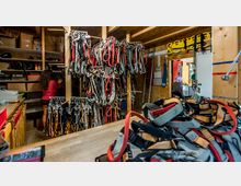Ein Lagerraum mit Regalen und zahlreichen hängenden Sicherheitsgurten in verschiedenen Farben. Im Hintergrund sortiert eine Person Gegenstände, und an den Wänden sind Leitern sowie weiteres Kletter- oder Rettungsequipment angebracht.