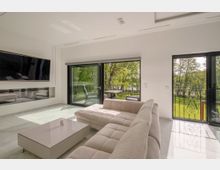 Modernes Wohnzimmer mit bodentiefen Glasschiebetüren, die auf einen Balkon mit Blick auf einen grünen Garten führen. Der Raum ist minimalistisch eingerichtet, mit einem großen beigen Sofa, einem flachen Couchtisch und einem wandmontierten Fernseher.