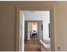 Innenansicht einer Wohnung mit Blick durch mehrere geöffnete Türen. Im Vordergrund ein heller Raum mit Holzboden, einem weißen Sofa und einer verzierten Türzarge; dahinter Essbereich mit Tisch, Stühlen und einem hängenden Kronleuchter.