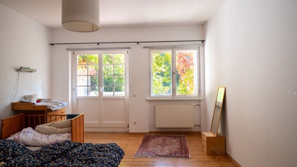 Helles, schlicht eingerichtetes Schlafzimmer mit Holzdielenboden, Doppelbett und kleiner Kommode. An der Stirnseite befinden sich eine verglaste Terrassentür und ein Fenster mit Blick ins Grün; dazu eine Deckenlampe, ein kleiner Teppich und ein Standspiegel an der Wand.