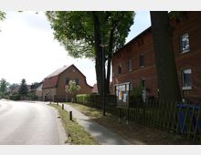 Straßenszene in einem Wohngebiet mit einem Gehweg, der an einer roten Backsteinmauer entlangführt. Auf der linken Seite stehen Häuser mit roten Dächern, ein Baum mit einer Straßenlaterne sowie ein Holzzaun und ein Anschlagbrett befinden sich im Vordergrund.